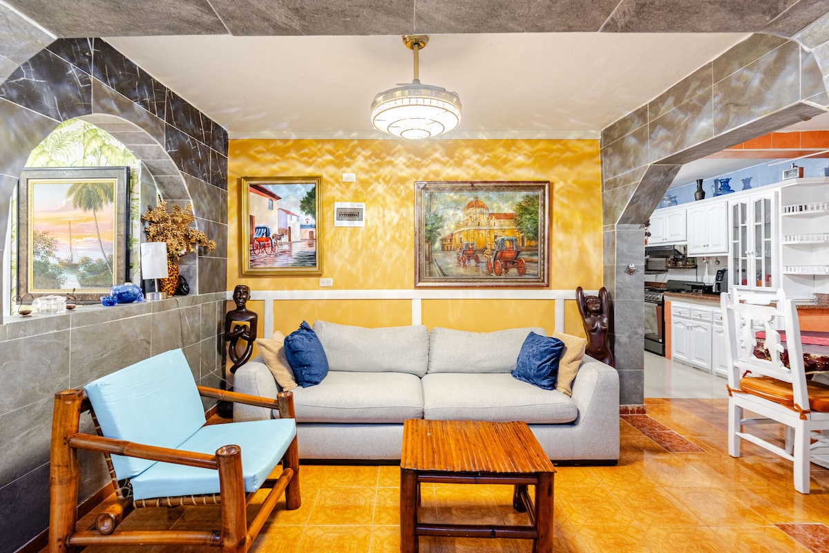 A spacious living area showcases a light grey sofa complemented by a wooden coffee table. A wooden armchair adds a pop of color, while paintings adorn the walls, reflecting local scenes. The architectural arches and soft lighting enhance the inviting ambiance.