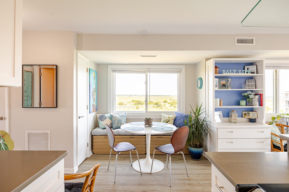 A bright and airy dining area features a round table surrounded by two modern chairs. A cozy nook with cushions offers a view of the marsh through large windows, complemented by decorative elements on the shelving unit and a potted plant.
