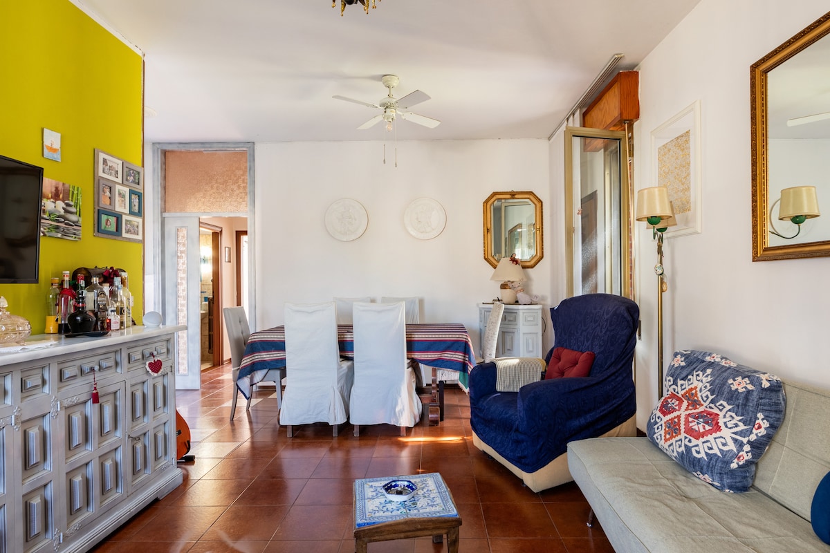 A bright living area features a blue armchair and a light-colored sofa. A dining table with white chairs is visible in the background, accompanied by a vibrant wall painted in yellow. Natural light flows through large windows, enhancing the open, welcoming atmosphere.