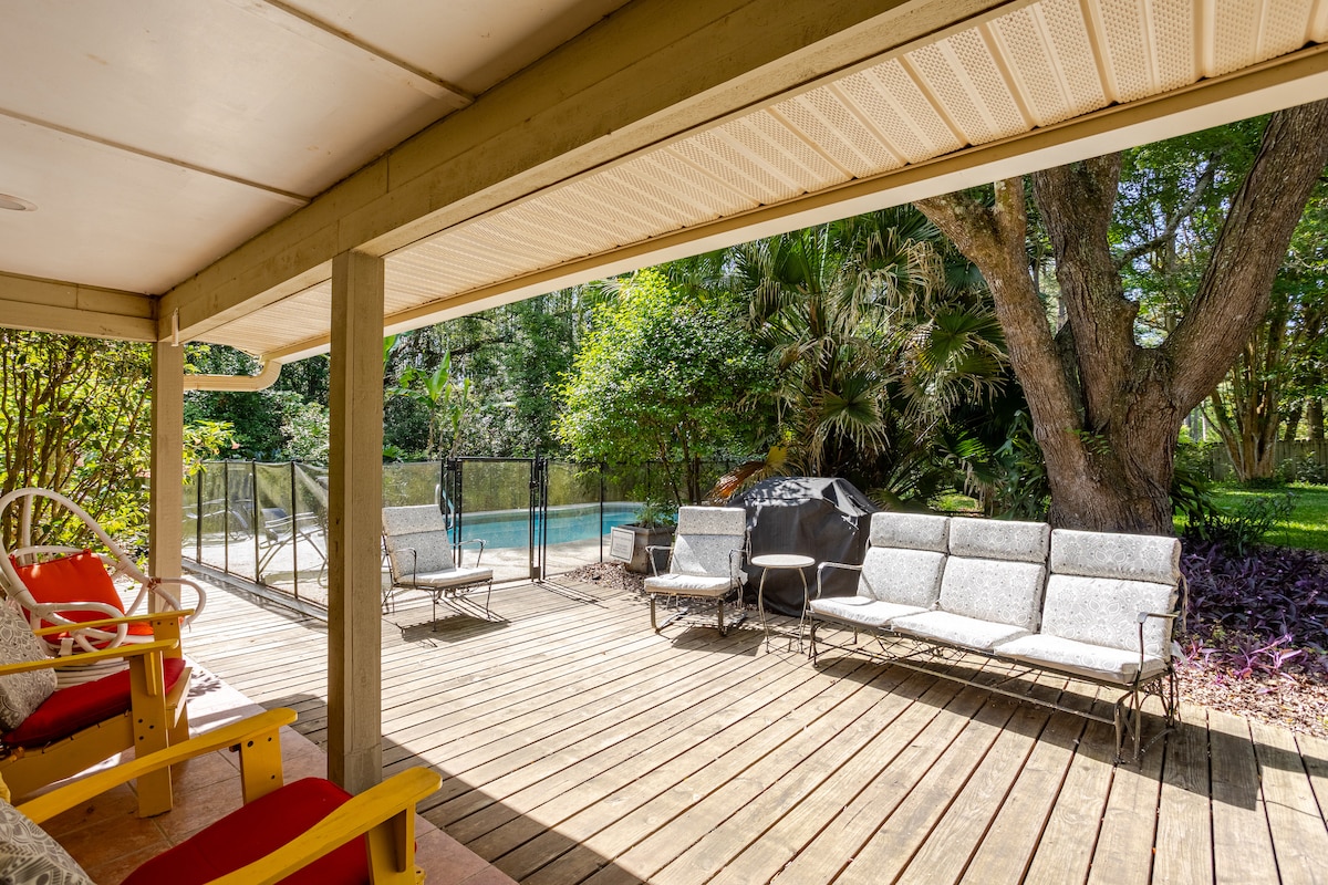 A spacious outdoor deck extends towards a serene pool area, enclosed for safety. Comfortable seating options, including lounge chairs and a side table, are arranged for relaxation, with lush greenery surrounding the space, creating a secluded atmosphere.