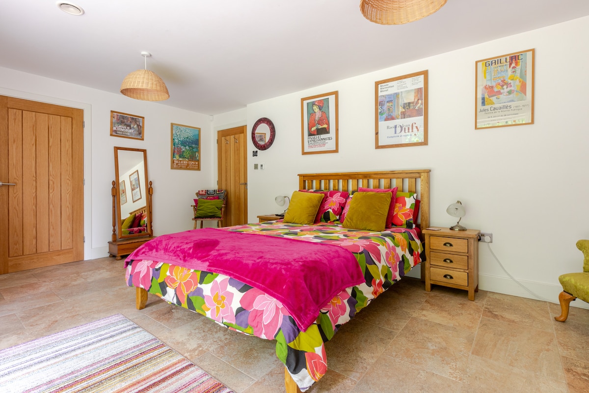 A bright bedroom features a wooden bed dressed in a colorful floral duvet with matching cushions. Artwork adorns the walls alongside a mirror and a cushioned chair sits nearby. Natural light enhances the spacious layout, complemented by stylish pendant lighting.