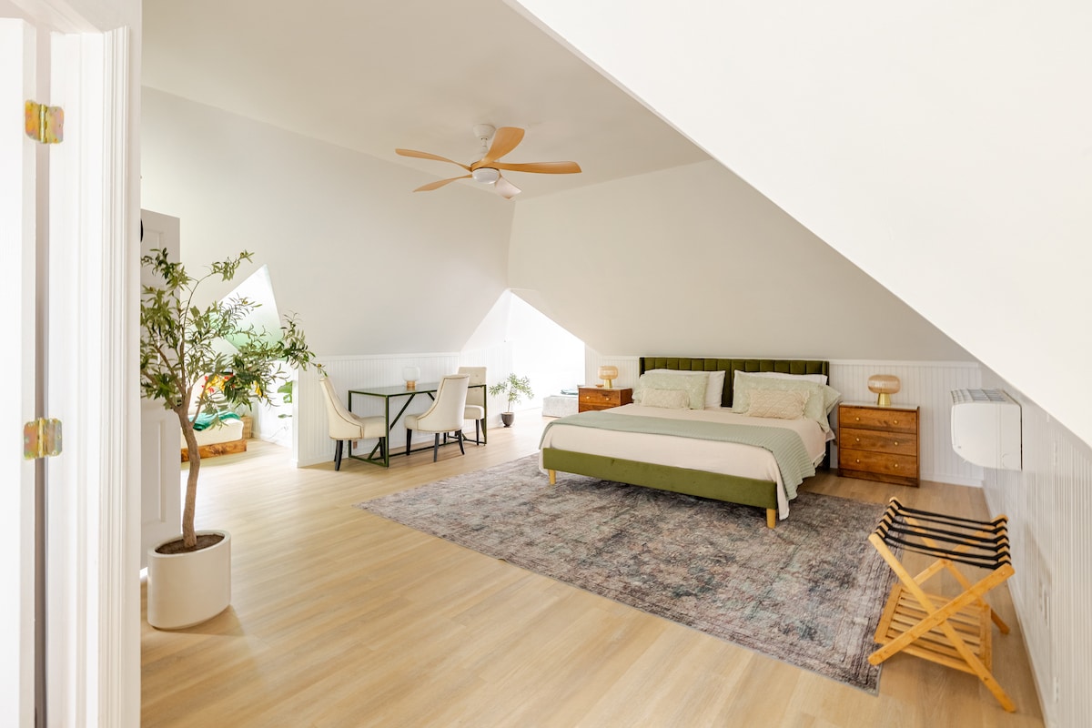 A spacious sleeping area is highlighted by a king bed with a green upholstered headboard. Natural light filters through triangular windows, creating a bright atmosphere. An area rug adds warmth to the wooden floor, while a small table and chairs are positioned at the far end.