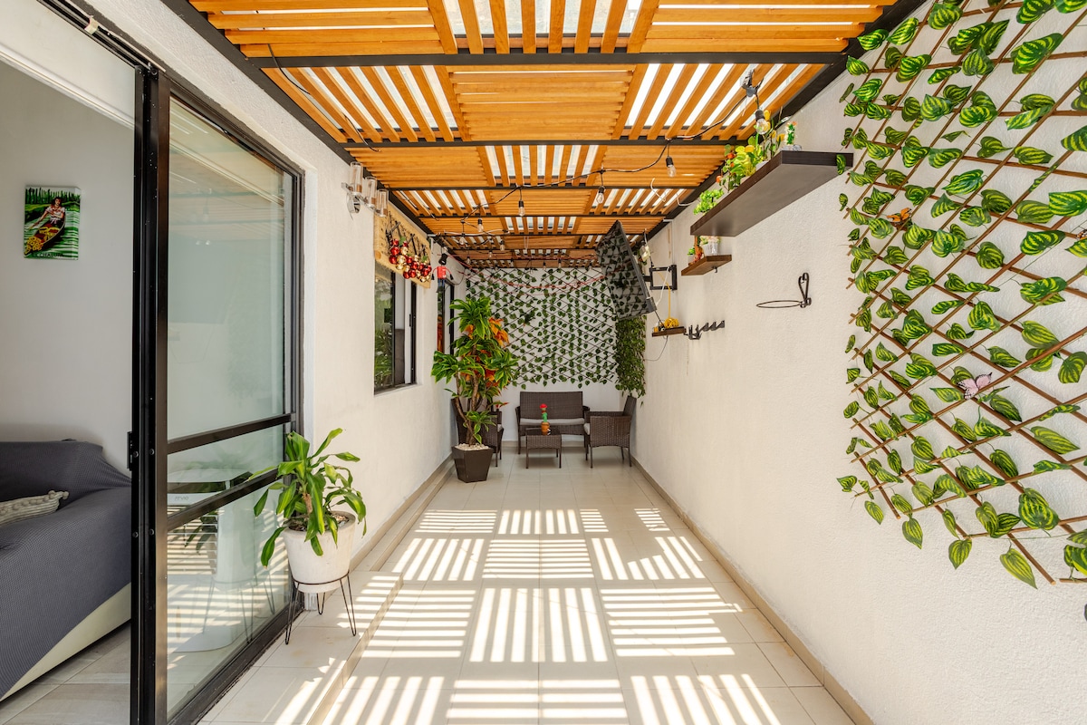 A covered terrace area features a wooden slatted roof allowing natural light to filter through. Lush green plants are arranged along the walls, while a seating arrangement of two chairs and a small table is present. The flooring is tiled, providing a clean and spacious appearance.
