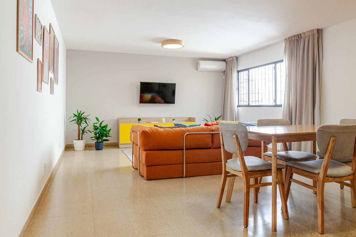 The living area features a bright and open layout with a comfortable orange sofa and a wooden dining table. A flat-screen TV is mounted on the wall, and large windows provide ample natural light. Potted plants add a touch of greenery to the space.