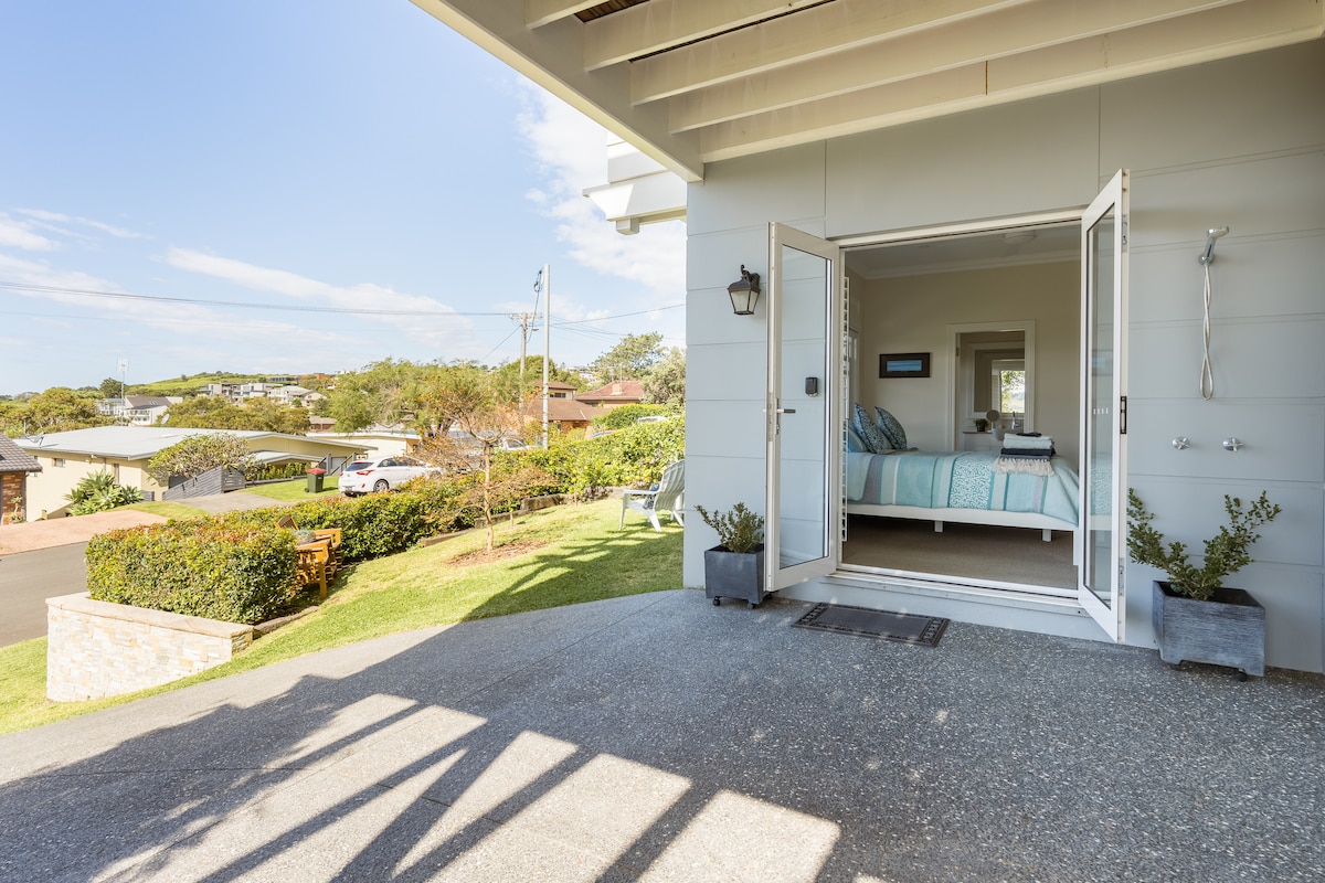 An inviting entry leads to a spacious bedroom with large doors open to the outdoors. The room features a king-sized bed with a light blue and white bedspread. Lush greenery and a view of the neighborhood are visible beyond the entryway.