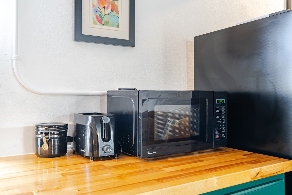 The kitchen countertop features a sleek black microwave, a toaster, and a container for storage. A modern refrigerator stands alongside, with a framed piece of artwork displayed on the wall above. The warm wooden surface enhances the functional space.