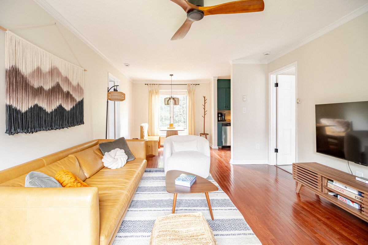The open-concept living room features a sleek leather sofa, a wooden coffee table, and a large television. Natural light flows in through the windows, highlighting the textured wall art and warm wood flooring. The space includes a ceiling fan and transitions to a dining area and kitchen.
