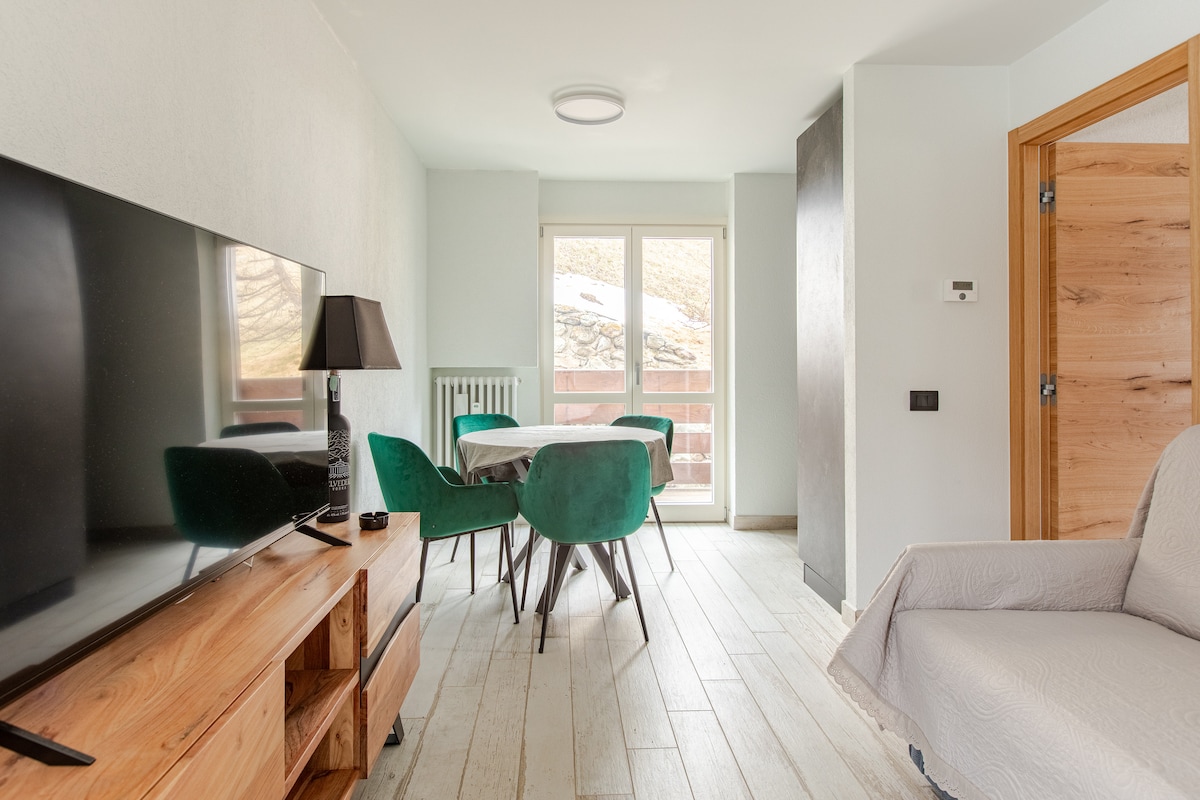 A light-filled living area is showcased, featuring a wooden television stand, a large screen TV, and a stylish lamp. A round dining table surrounded by green chairs is positioned near a sliding glass door that opens to a balcony, bringing the outdoors in.
