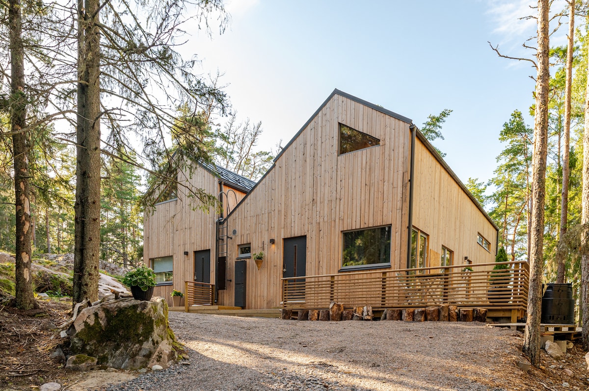 The exterior of a modern wooden cottage is surrounded by trees, showcasing a combination of large windows and a natural wooden facade. A spacious deck is visible in front, with an inviting entryway leading into the cabin.