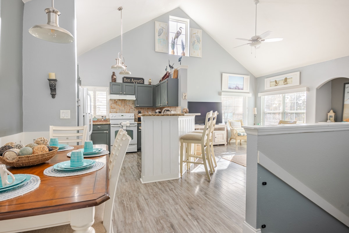 An open-concept kitchen and dining area is featured, showcasing a wooden dining table set for four with turquoise dishes. A well-equipped kitchen with dark cabinetry and modern appliances is visible in the background, while natural light filters through nearby windows, illuminating the space.