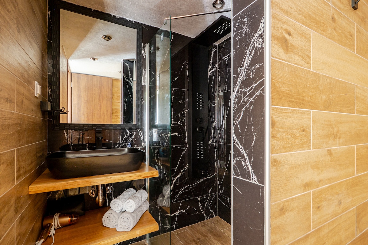 A modern bathroom features stylish wooden and marble accents. A spacious walk-in shower is equipped with multiple showerheads, while a sleek black sink rests on a wooden shelf. Freshly rolled towels are neatly arranged on the lower shelf, contributing to an organized appearance.