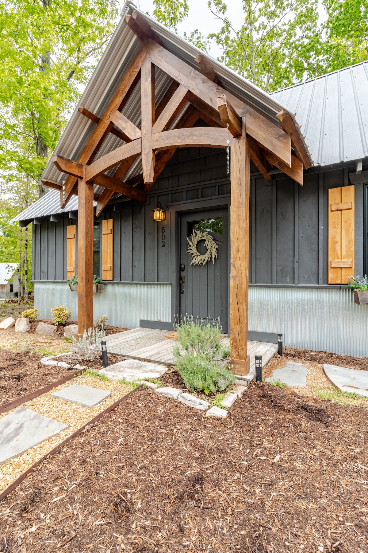 The entrance to the cabin is framed by a large wooden overhang supported by exposed beams. The door is accented by a circular wreath, while decorative shutters flank the windows. Mulch and minimal landscaping surround the path leading to the entrance, creating a welcoming entryway.