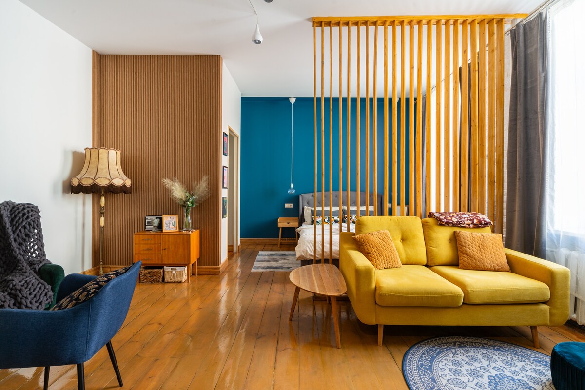 A living area features a yellow sectional sofa, complemented by a blue armchair and a modern wooden coffee table. A stylish lamp stands beside a wooden cabinet. The space is divided by a wooden slatted screen, leading to a cozy sleeping area with a turquoise wall.