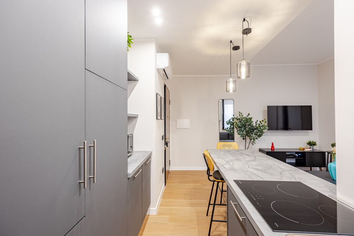 An open kitchen space features sleek grey cabinetry and a marble countertop. A modern induction cooktop is visible, complemented by stylish pendant lighting overhead. The room includes a dining area with yellow chairs, and a large window allows natural light to fill the space.