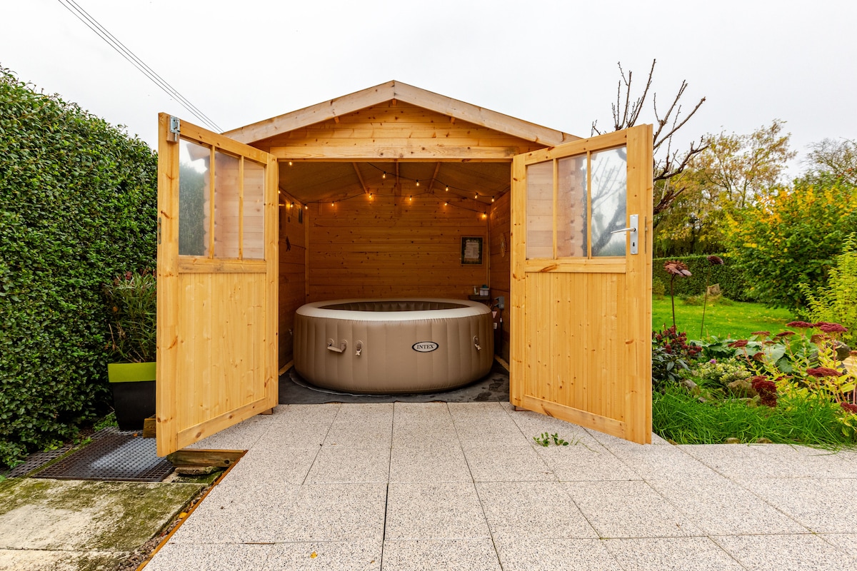 A wooden chalet features a spacious hot tub, framed by double doors that invite natural light. Soft lighting enhances the cozy atmosphere within. The surrounding garden presents lush greenery and colorful plants, creating a serene outdoor space.