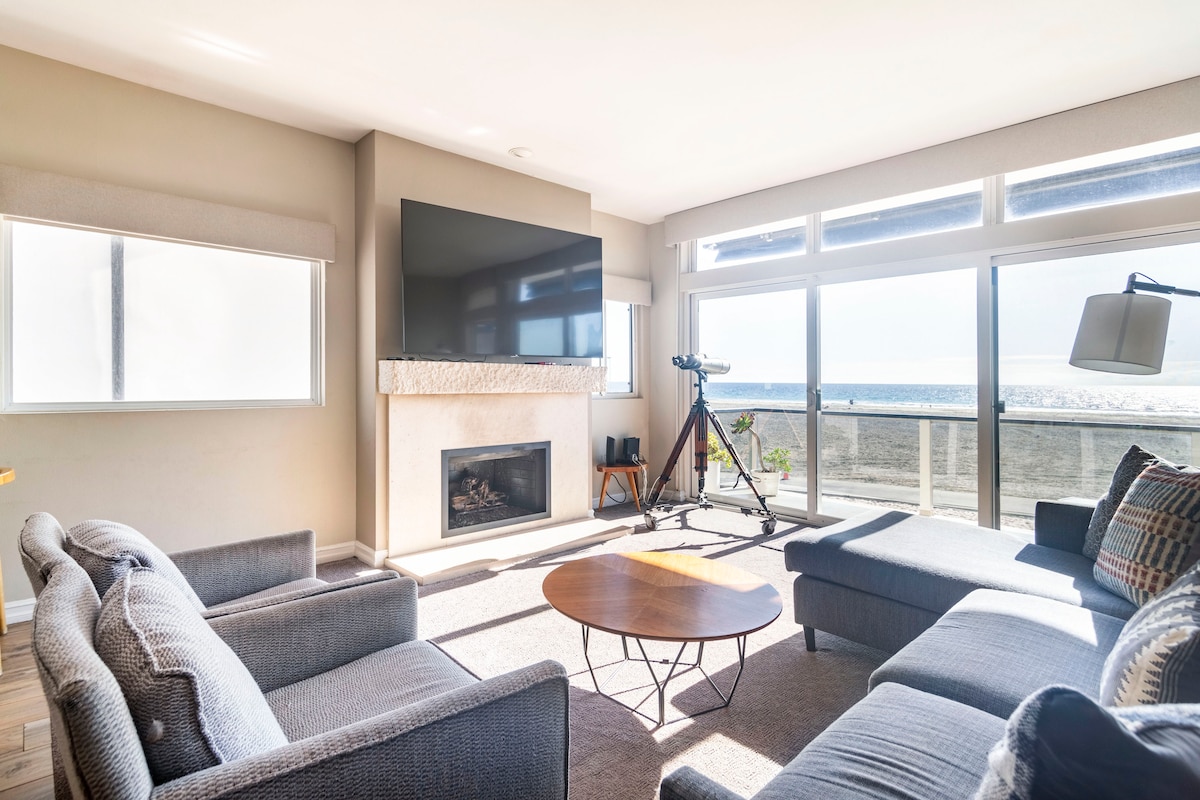 A living room area is furnished with comfortable sofas and a round table. Large windows allow for ample natural light, framing spectacular ocean views. A flat-screen TV is mounted above a fireplace, while a telescope is positioned beside the window for seaside observation.