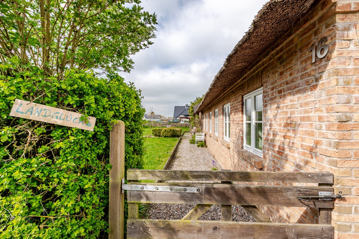 A charming entrance is framed by lush greenery, leading to a rustic thatched-roof building. The sign 'Landglück' is positioned on a wooden gate, welcoming guests. Several windows allow natural light to enter the building, creating an inviting atmosphere throughout the exterior.