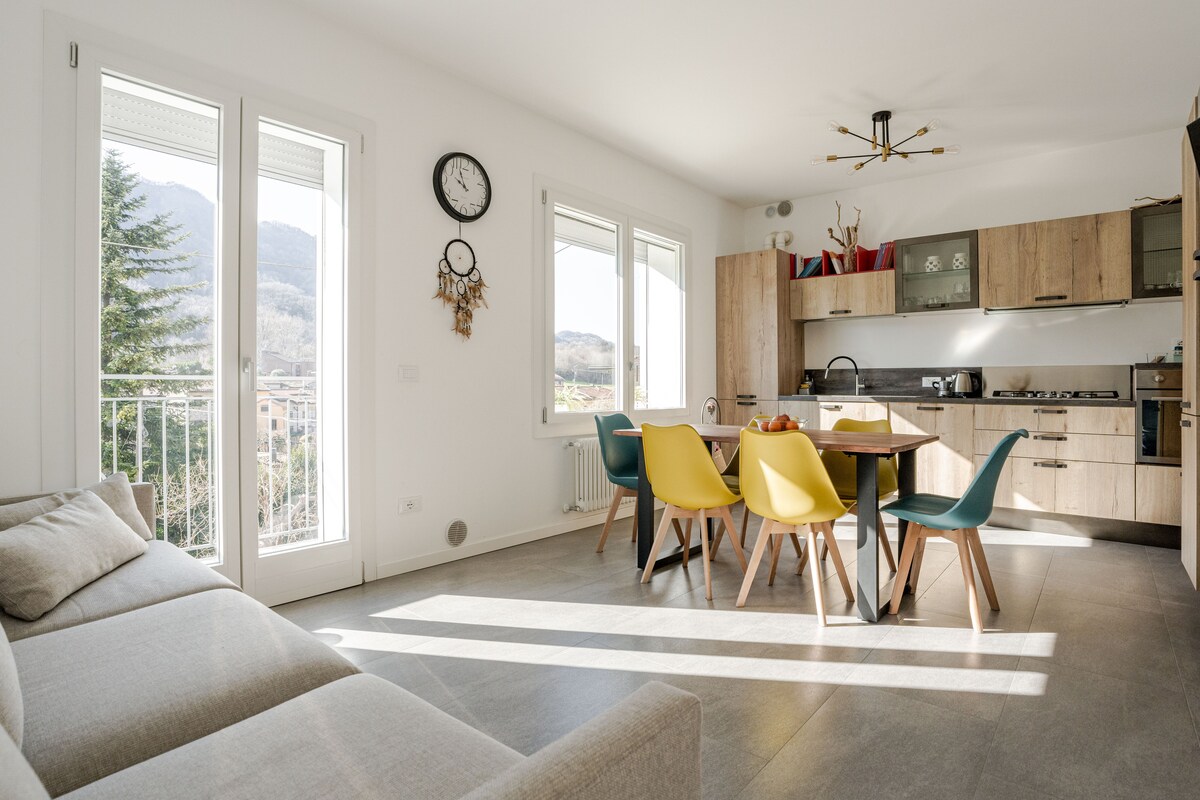A bright dining area features a wooden table surrounded by six chairs in various colors. Large windows allow natural light to fill the space and provide views of the mountains. A cozy sofa is placed nearby, enhancing the inviting atmosphere of the room.