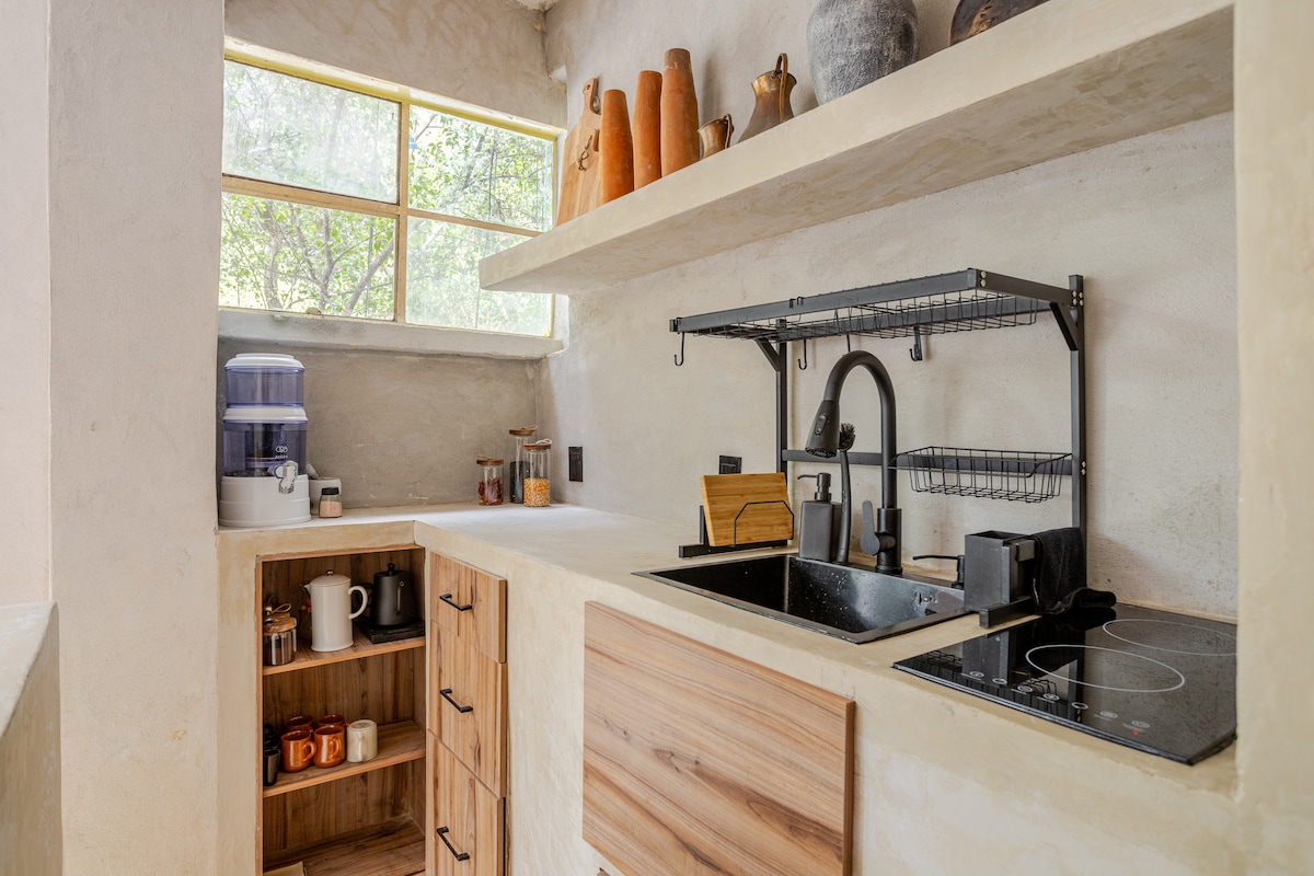 A modern kitchen features natural materials with a light and neutral color palette. Open shelves display clay pots and decorative items. A sleek sink with a black faucet is positioned prominently alongside an induction cooktop, while a wooden storage unit holds various kitchen essentials.