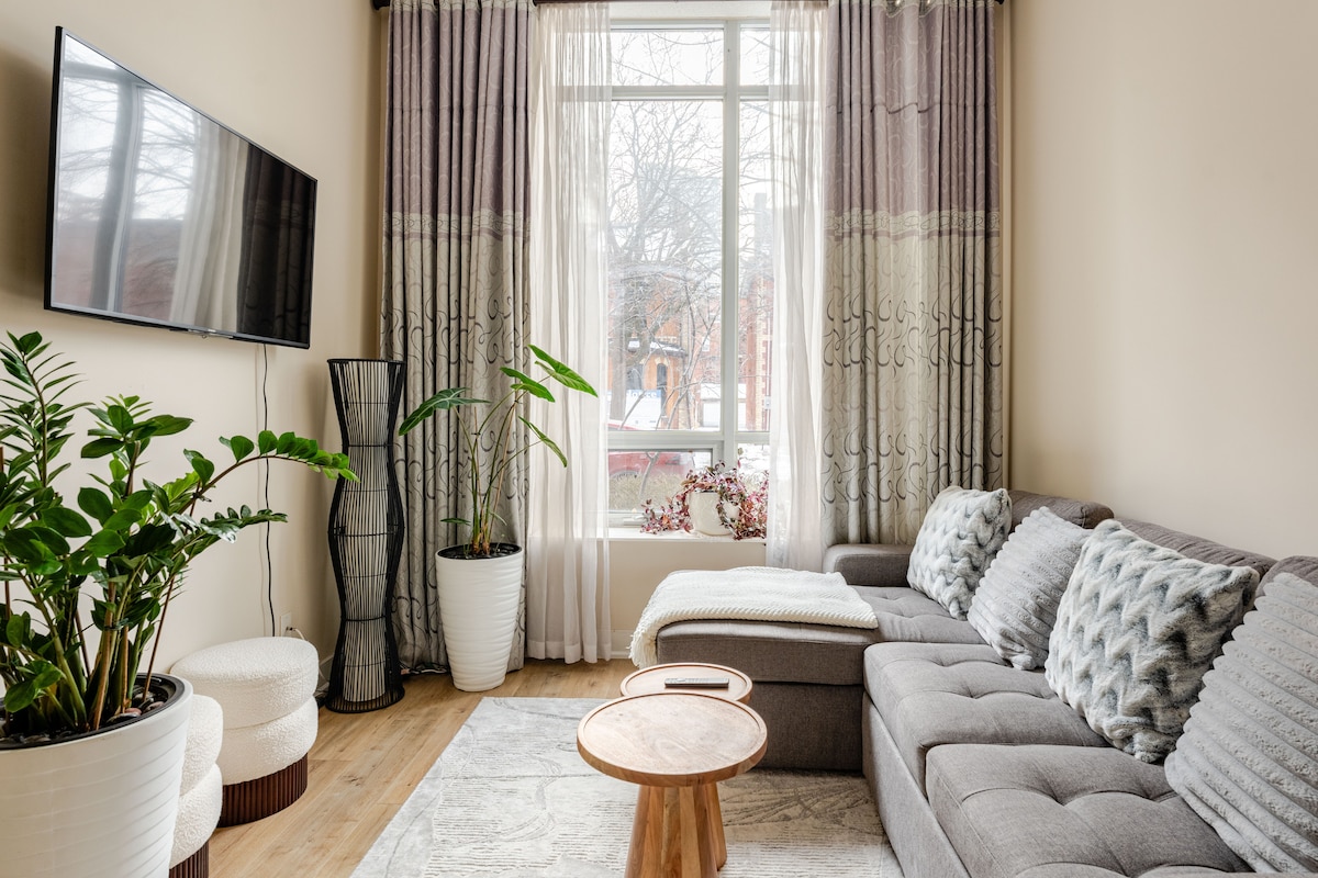 A cozy living area features a spacious grey sectional sofa and a wooden coffee table. Natural light floods in through a large window dressed with soft, patterned curtains. Two potted plants add a touch of greenery, enhancing the inviting atmosphere.