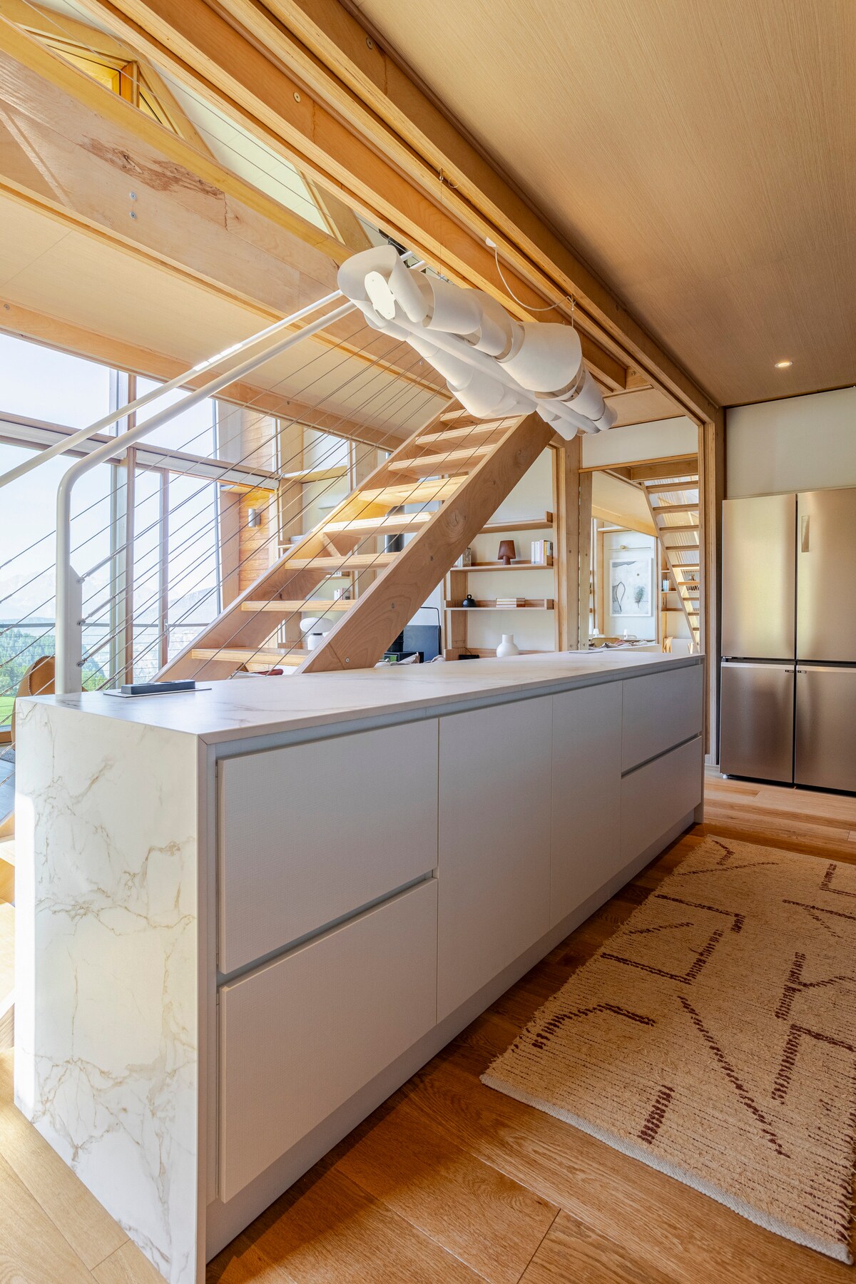 A modern kitchen is displayed, featuring a central island with a marble countertop. Natural light is captured through large windows, enhancing the bright wooden interior. An elegant staircase with wooden steps leads to the upper level, while a woven rug adds texture to the floor.