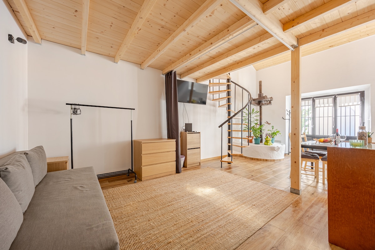 A spacious living area is showcased with natural wood beams and flooring. A comfortable sofa is placed near a sleek television mounted on the wall. A staircase leads to an upper level, while a dining area with chairs and a table is visible in the background.