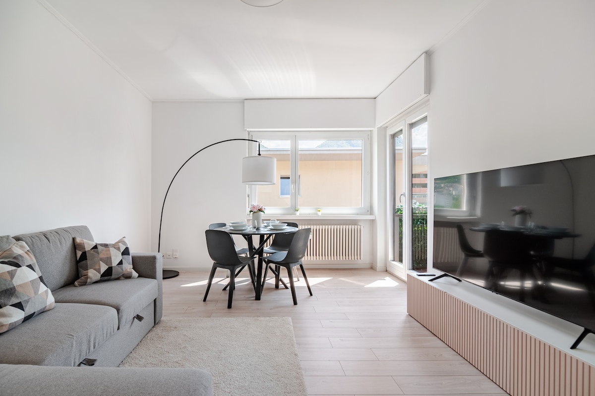 A bright living area features a comfortable gray sofa adorned with patterned cushions. A round dining table with black chairs is situated beside a large window, allowing natural light to fill the space. A flat-screen television is positioned against a wooden panel, enhancing the clean lines of the room.