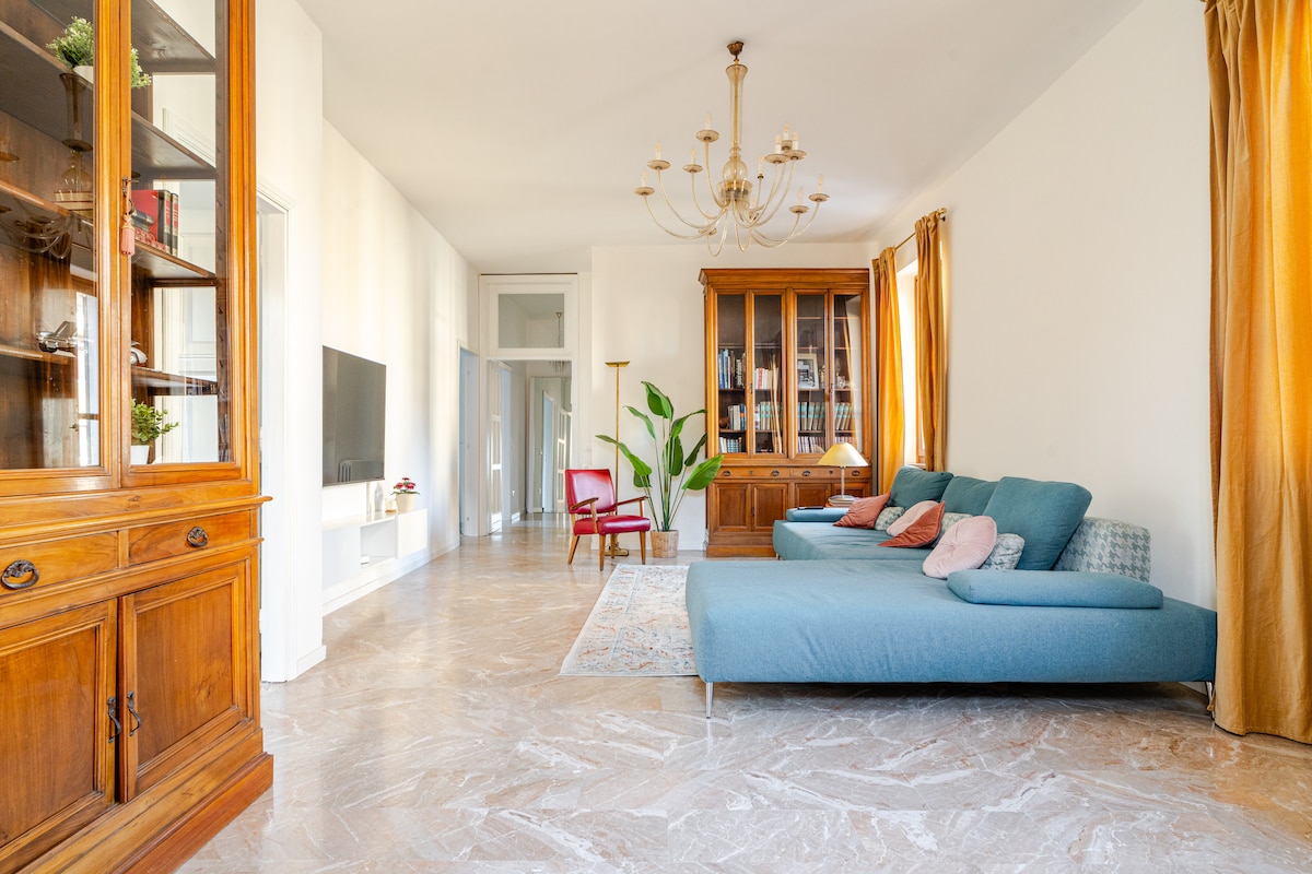 A spacious living room features a comfortable blue sofa with multiple cushions. Natural light fills the area, enhancing the wooden cabinetry and light-colored walls. A large area rug sits on patterned flooring, complemented by an elegant light fixture and tall drapes framing the windows.