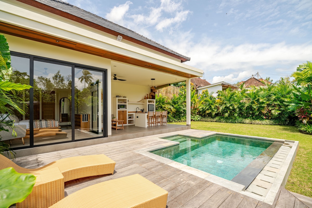 An outdoor area features a small private pool adjacent to a wooden deck with two lounge chairs. The open living space is visible through expansive glass doors, providing views of the kitchen and dining area. Lush greenery surrounds the villa, enhancing the tranquil setting.