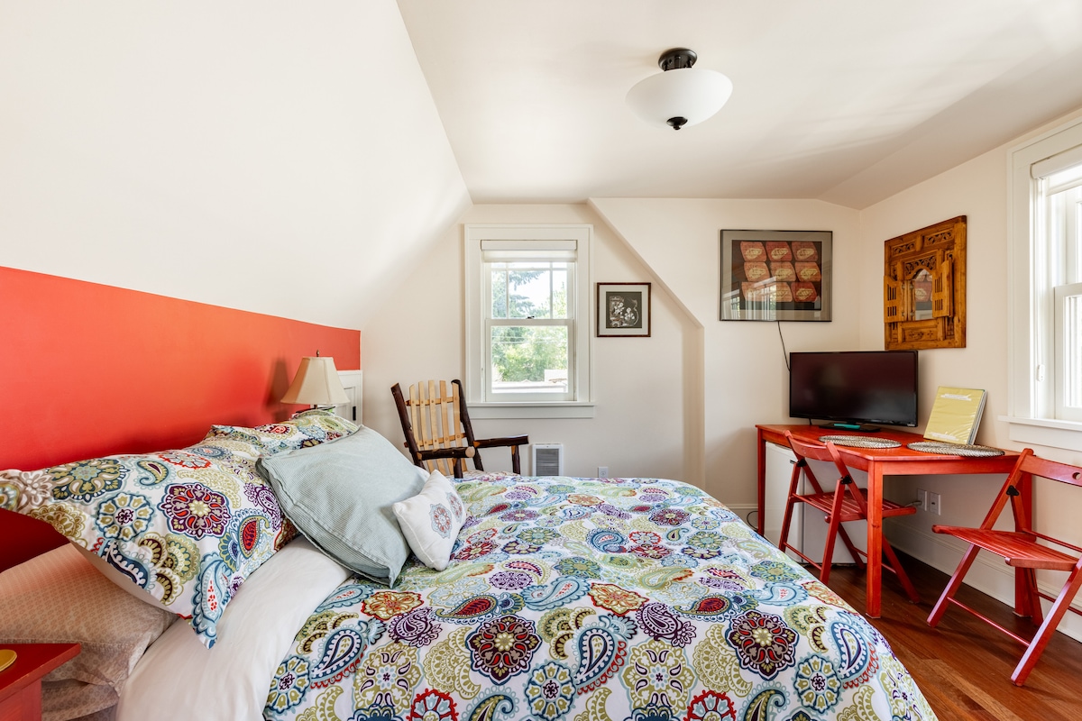 A bright bedroom features colorful patterned bedding on a double bed. A small desk with red chairs is positioned near a window. An armchair sits in one corner, and framed art adorns the walls. Natural light enhances the cheerful ambiance.