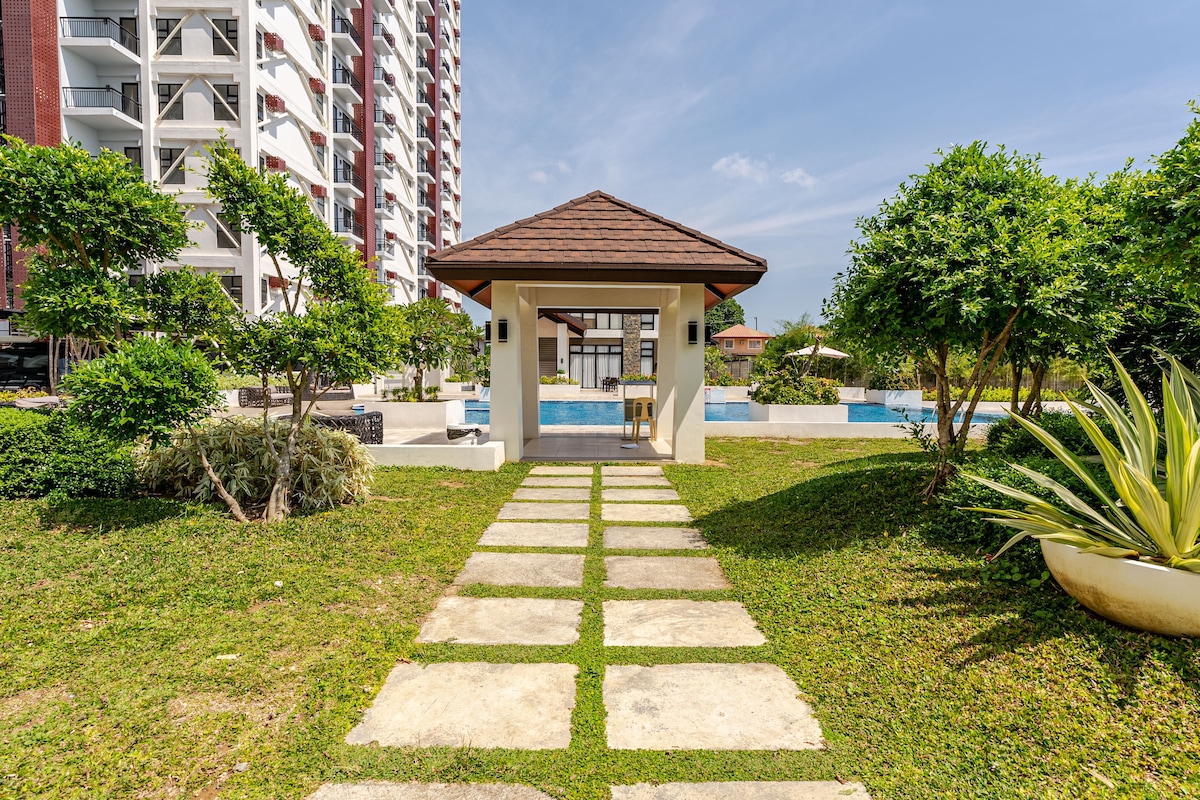 A pathway of stone tiles leads through a well-maintained grassy area towards a covered gazebo. The gazebo is set against a backdrop of a modern building and features greenery and a pool visible in the distance under clear blue skies.
