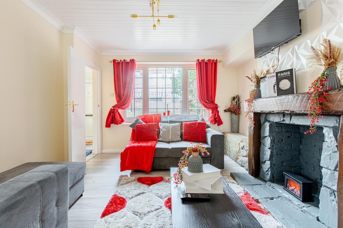 A spacious living room features a gray sectional sofa adorned with red and white cushions. A decorative fireplace sits beneath a wall-mounted television. Red curtains frame the window, allowing soft natural light to illuminate the room. A coffee table with books and a floral arrangement completes the space.