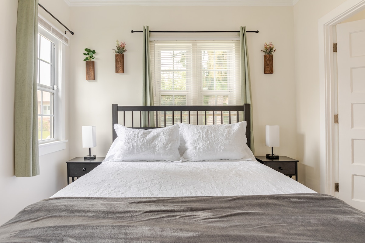 A bright sleeping area is furnished with a spacious bed, topped with a white quilt and decorative pillows. Nightstands are placed on either side, each adorned with a sleek lamp. Natural light filters through window treatments, enhancing the airy feel of the room.