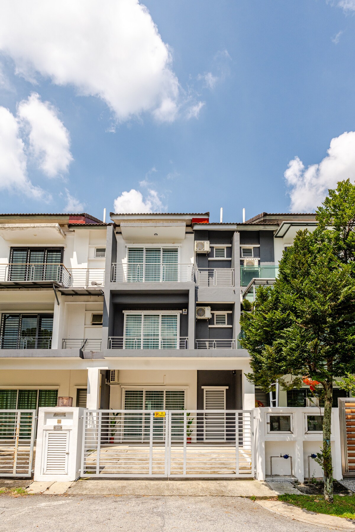 The exterior of the multi-story house is shown under a clear sky. A gated entrance leads to a wooden deck, framed by greenery. Large windows and balconies are highlighted, showcasing contemporary architectural features with a mix of light and dark-colored façades.