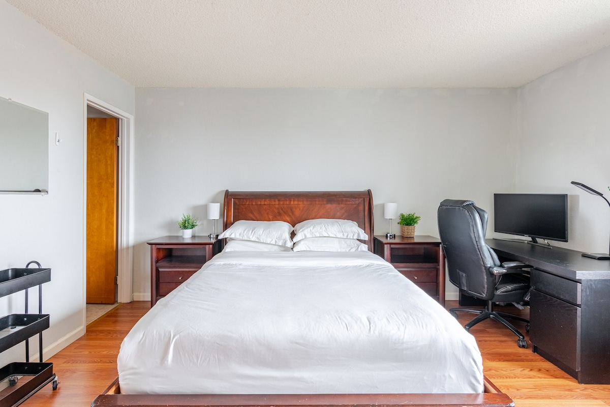A spacious bedroom contains a large bed with crisp white bedding. Nightstands are placed on either side, adorned with small plants. A desk area with a computer monitor and an ergonomic chair is nearby, while a mirror adds to the room's functionality. Natural light filters in from a window.