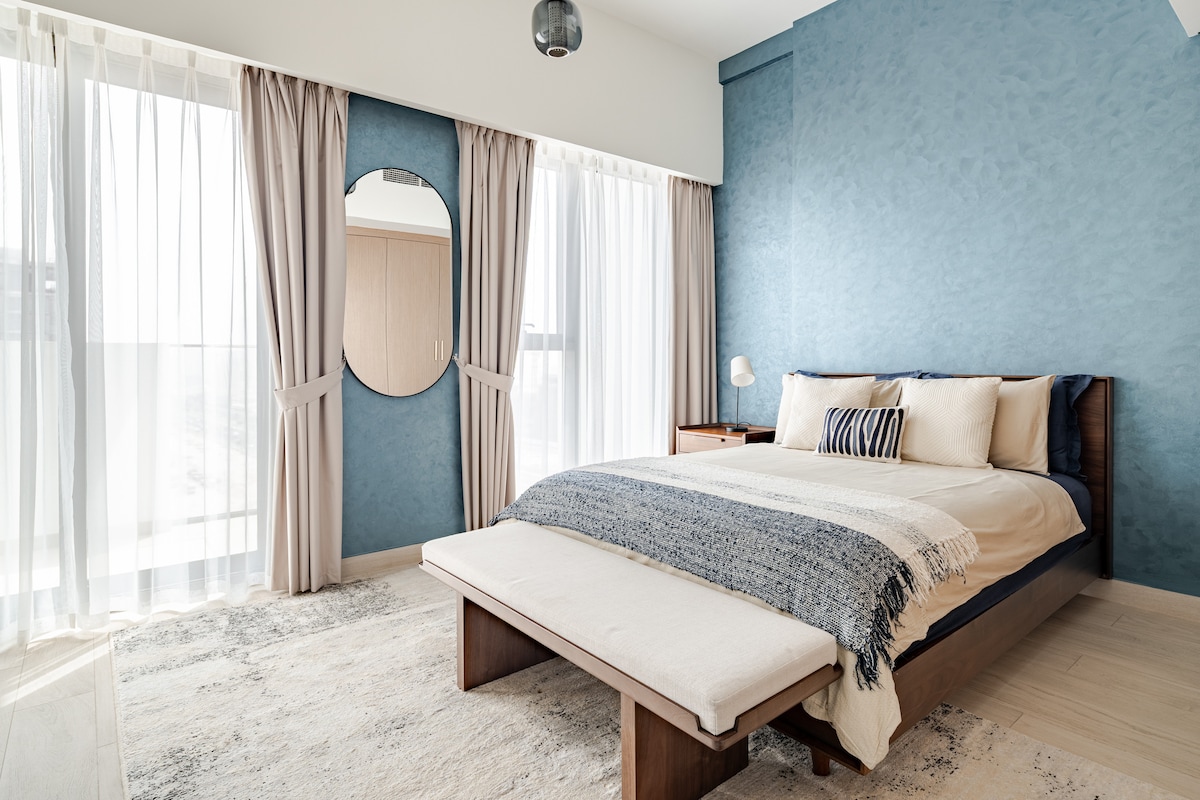 A serene sleeping area features a spacious bed dressed in soft linens and decorative pillows. Blue walls complement the natural light streaming in through floor-to-ceiling curtains, while a round mirror and nightstand add functional elegance. A plush rug enhances the cozy atmosphere.
