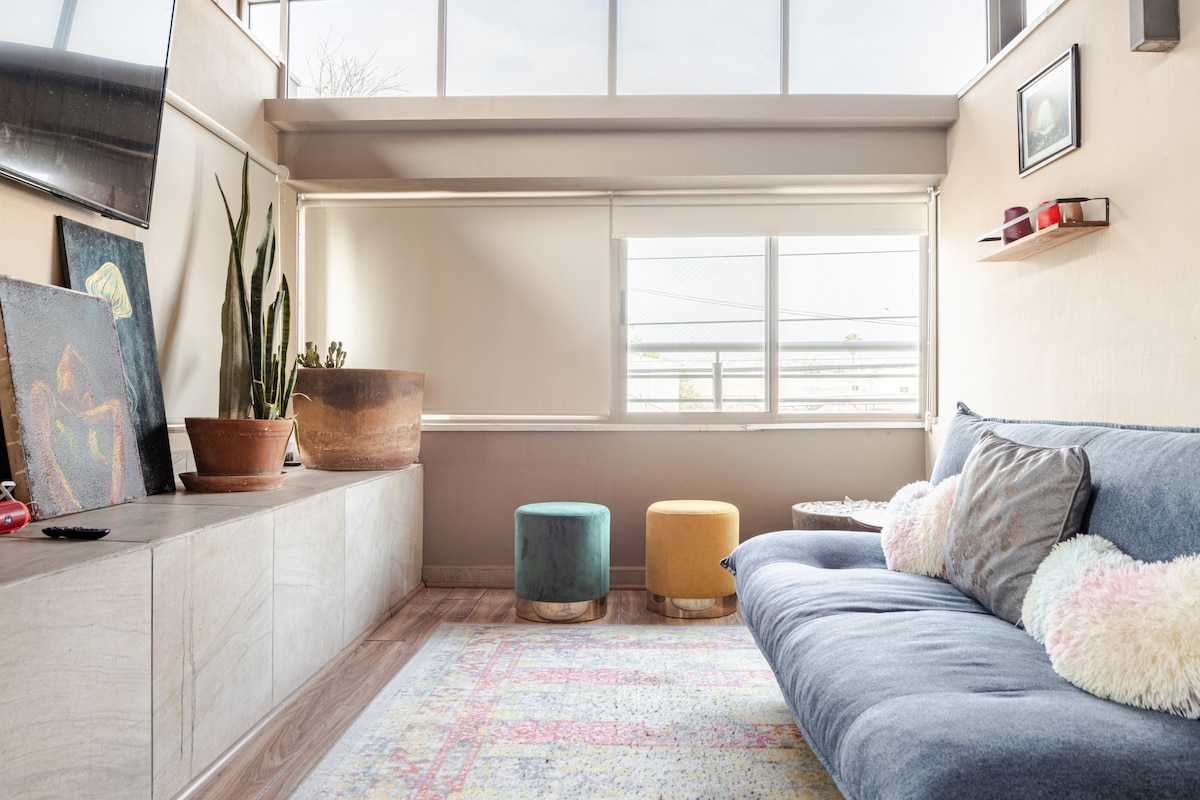 A comfortable living area features a spacious sofa adorned with plush cushions. Two colorful ottomans sit nearby, and natural light filters in through large windows, illuminating the room. Decorative shelves display art, while a potted plant adds a touch of greenery.