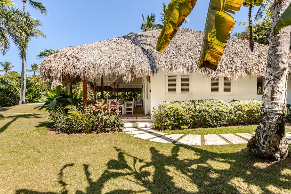 A charming villa with a thatched roof is nestled among tropical greenery. Lush plants surround a stone pathway leading to a covered porch, which features white wooden furniture. The pristine lawn is dotted with palm trees, creating a serene and inviting setting.