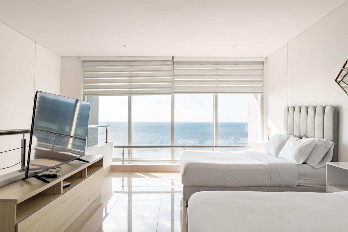 A spacious bedroom features two beds with crisp white linens, complemented by light-colored furniture. A large television is positioned on a cabinet. Floor-to-ceiling windows provide an unobstructed view of the ocean, with shades drawn halfway to allow natural light into the space.