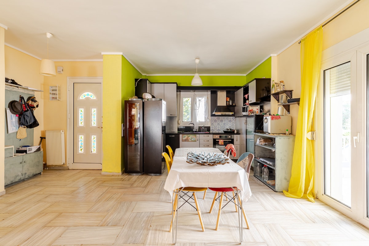 A spacious kitchen area features a dining table at its center, surrounded by colorful chairs. Sleek black cabinets and appliances are present against lively green and yellow walls. Natural light enters through large glass doors, illuminating the room.
