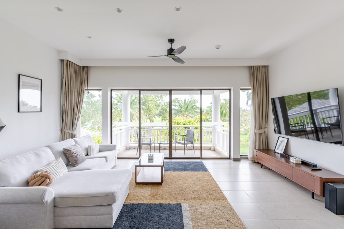 A spacious living area features a light-colored sectional sofa and a modern coffee table. Large glass doors provide access to a terrace, revealing views of the surrounding greenery. A flat-screen TV is mounted on a wooden console, complementing the room's neutral tones.