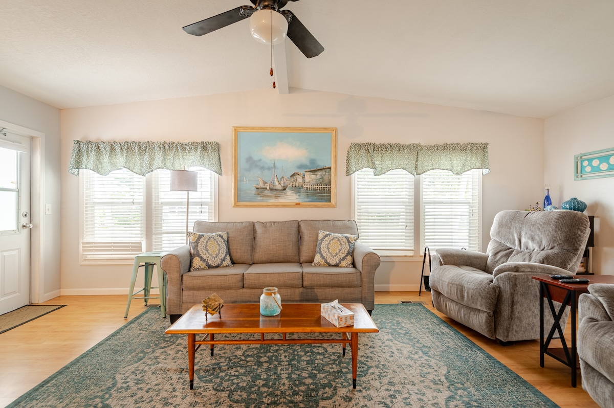 A bright living area features a comfortable gray sofa and a plush, reclining armchair. Natural light streams through multiple windows, complemented by patterned curtains. A rectangular wooden coffee table is centrally placed, and a large painting above the sofa adds a decorative touch to the wall.