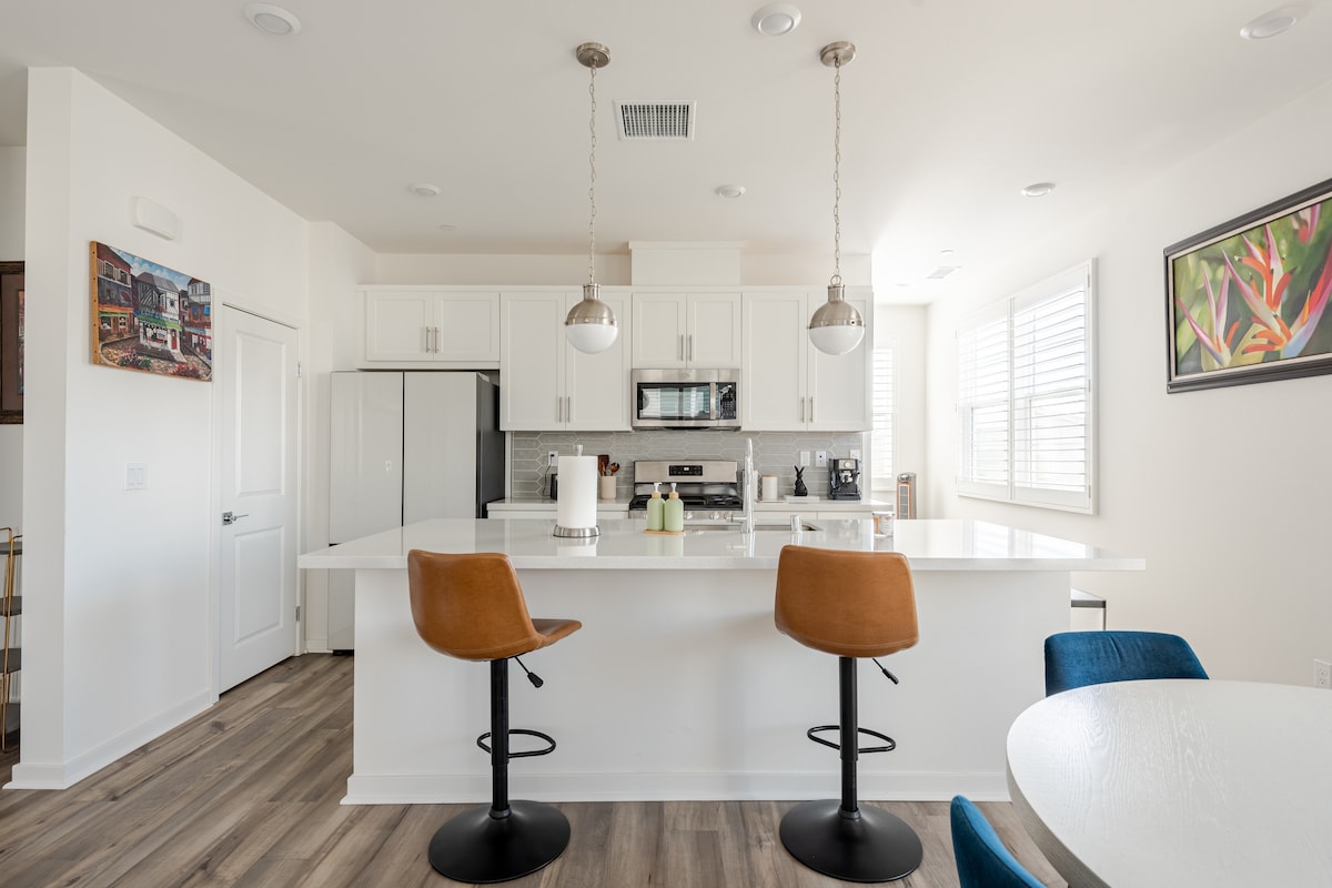 A modern kitchen is presented with light cabinetry and a white countertop. Two high-backed stools are placed at the kitchen island, while a dining table sits nearby. Natural light fills the space through window shutters, highlighting the appliances and decor.