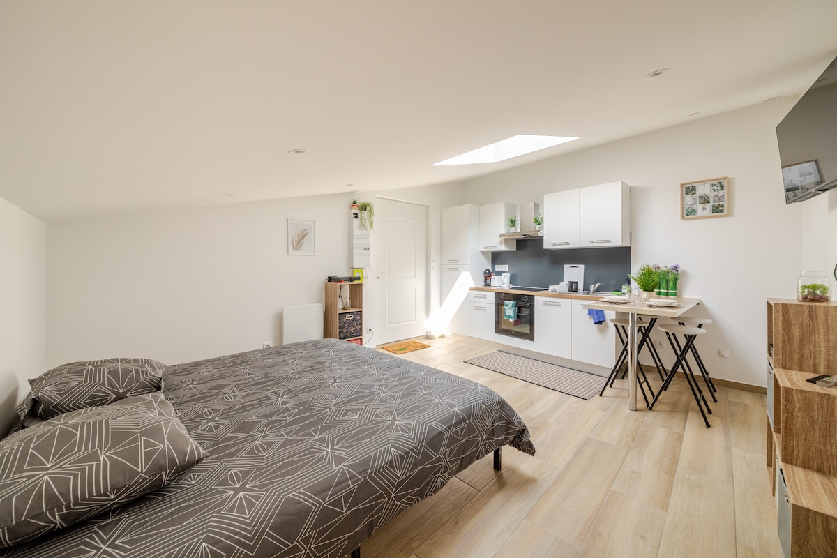 An airy studio apartment is presented, featuring a king-size bed with a geometric patterned duvet cover. A modern kitchenette with white cabinetry is visible, along with a small dining table and chairs. Large windows allow natural light to brighten the space.
