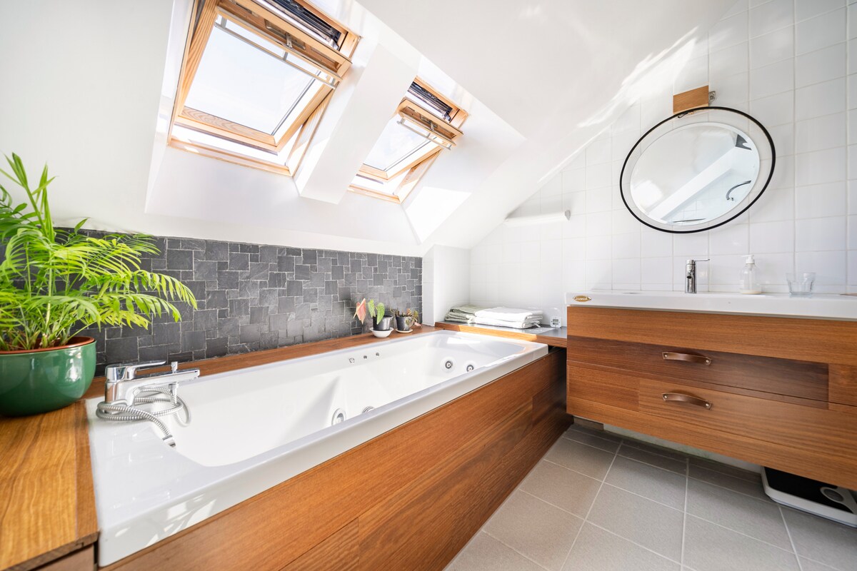 A well-appointed bathroom features a spacious bathtub with a wood panel surround, complemented by a large mirror above a wooden vanity. Natural light enters through two skylights, and fresh greenery is placed in a potted plant, enhancing the serene atmosphere.