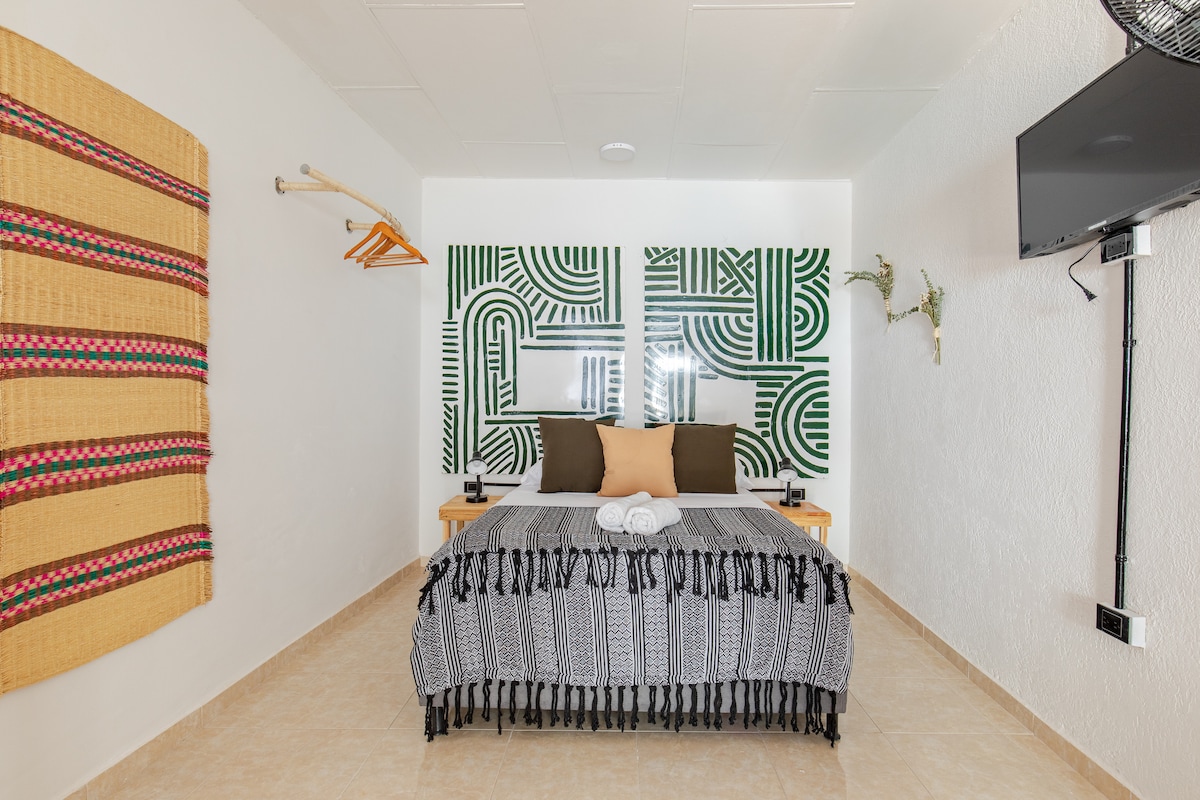 A well-appointed sleeping area features a spacious bed adorned with a patterned black and white blanket. Two decorative wall panels with green geometric designs complement the neutral-toned walls. A flat-screen TV is mounted nearby, and wooden shelves are positioned for storage.