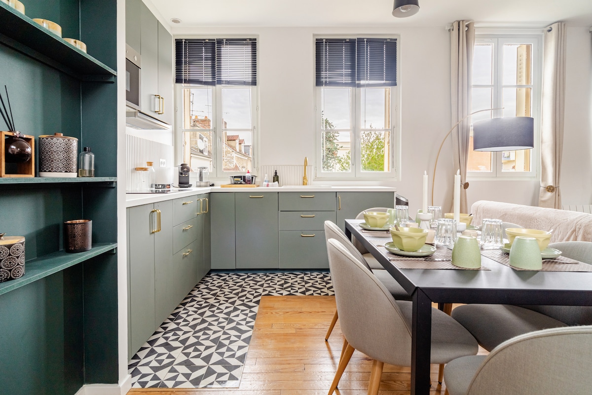 A modern kitchen and dining area feature sleek cabinetry in muted green tones, paired with a geometric-patterned rug. The dining table, set for a meal, is surrounded by elegant gray chairs. Natural light fills the space through large windows, creating a bright and functional atmosphere.