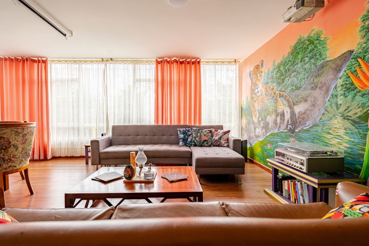 Een kamer met een eclectische en levendige stijl, vol natuurlijk licht en unieke details: tropische muurschildering, comfortabele banken, boeken, projector en surround sound. Een moderne en warme ruimte, perfect om te lezen, te ontspannen of van je favoriete film te genieten.