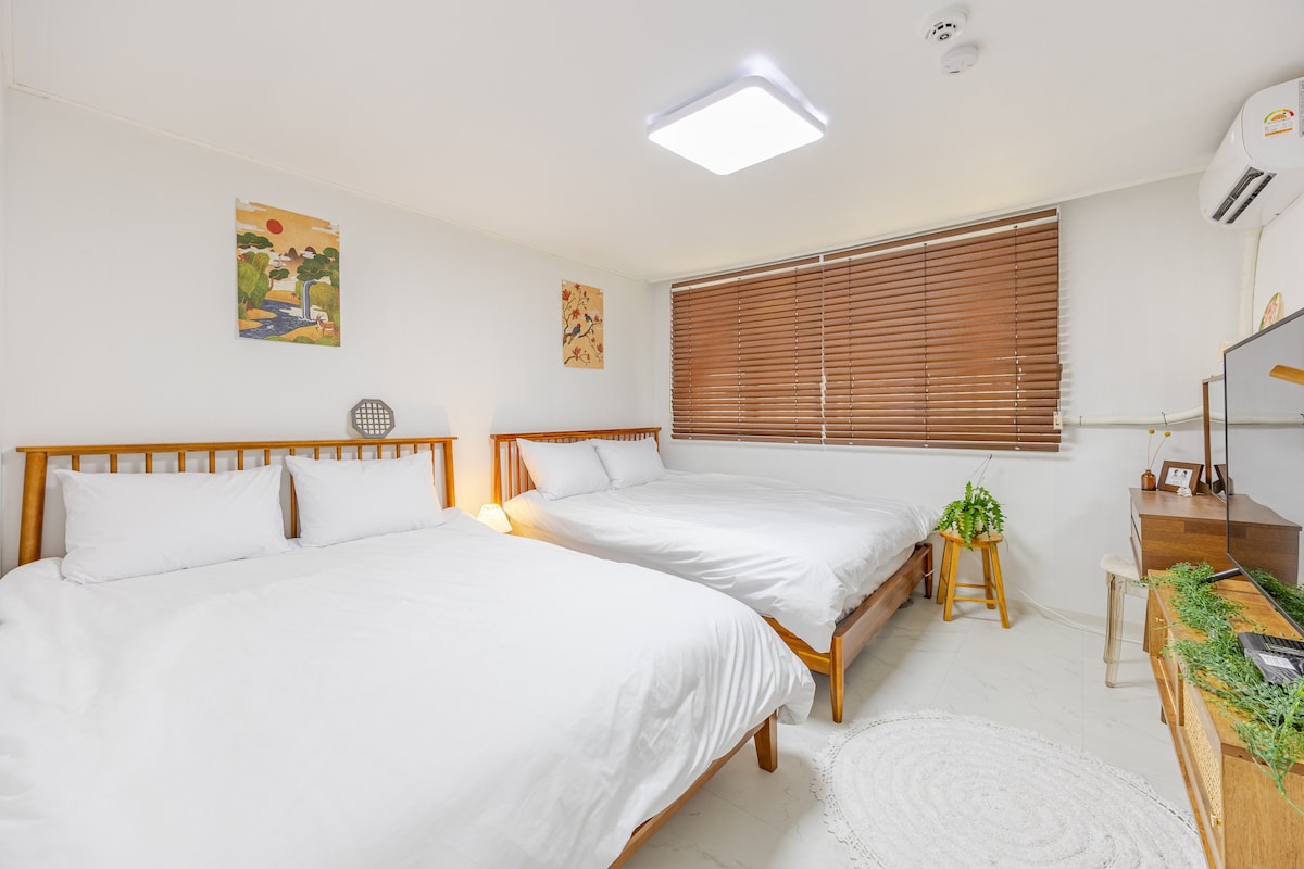 The bedroom features two spacious queen beds, each dressed in crisp white bedding. Natural light enters through a large window with wooden blinds. Minimalist decor, including wall art and a small plant, enhances the serene atmosphere of the space.