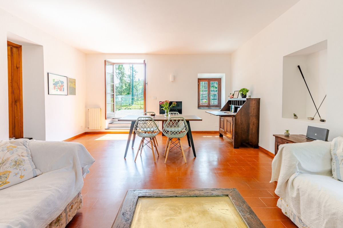 A spacious living area features two cozy sofas and a dining table surrounded by woven chairs. Natural light enters through the large window and door, illuminating the terracotta floors. A wooden cabinet and a small decorative item contribute to the room's inviting ambiance.